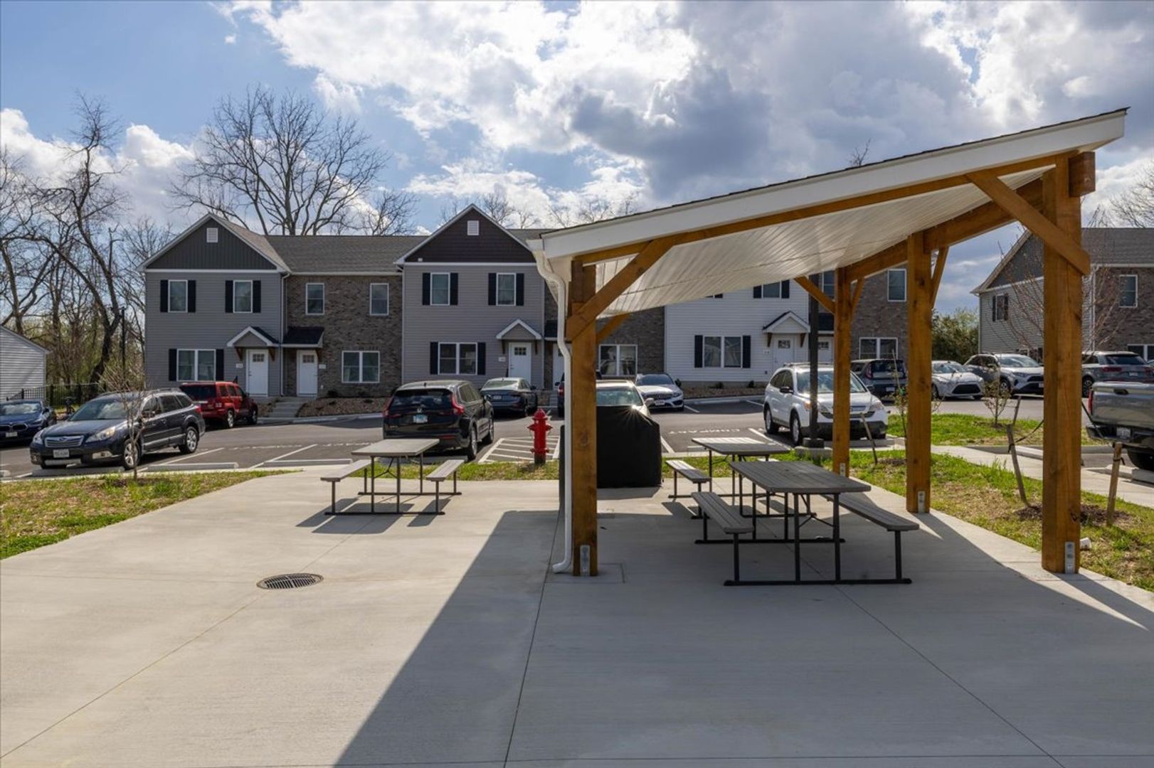 A concrete park area with picnic tables and a wooden shelter. Townhouses and cars are in the background.