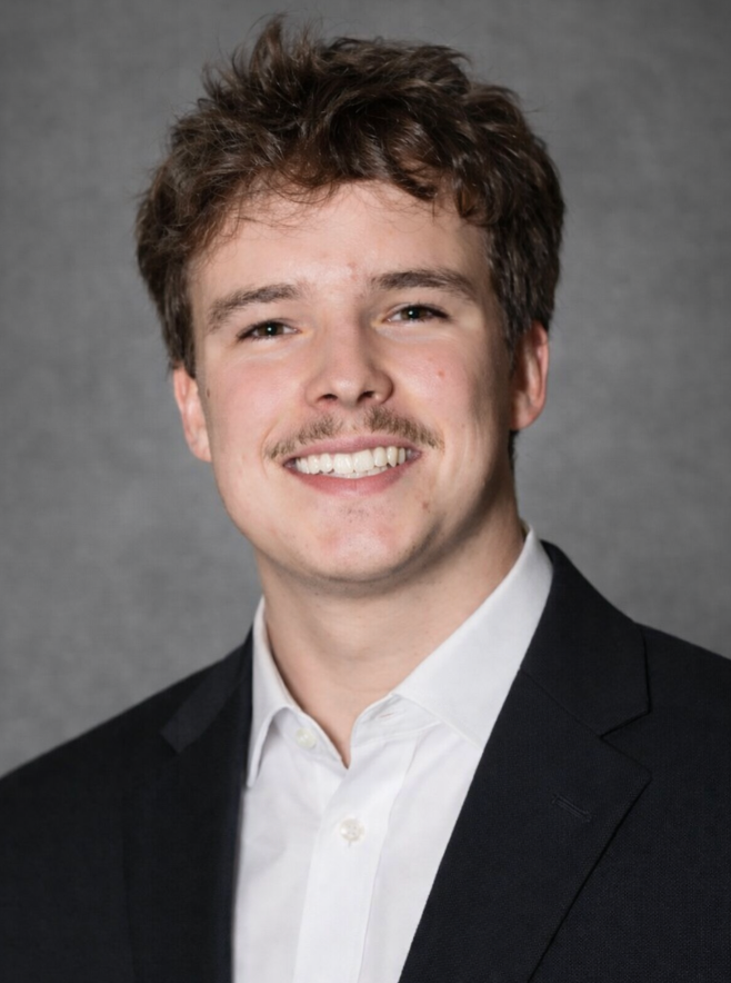 Man with dark curly hair and a mustache smiling, wearing a dark blue collared shirt.