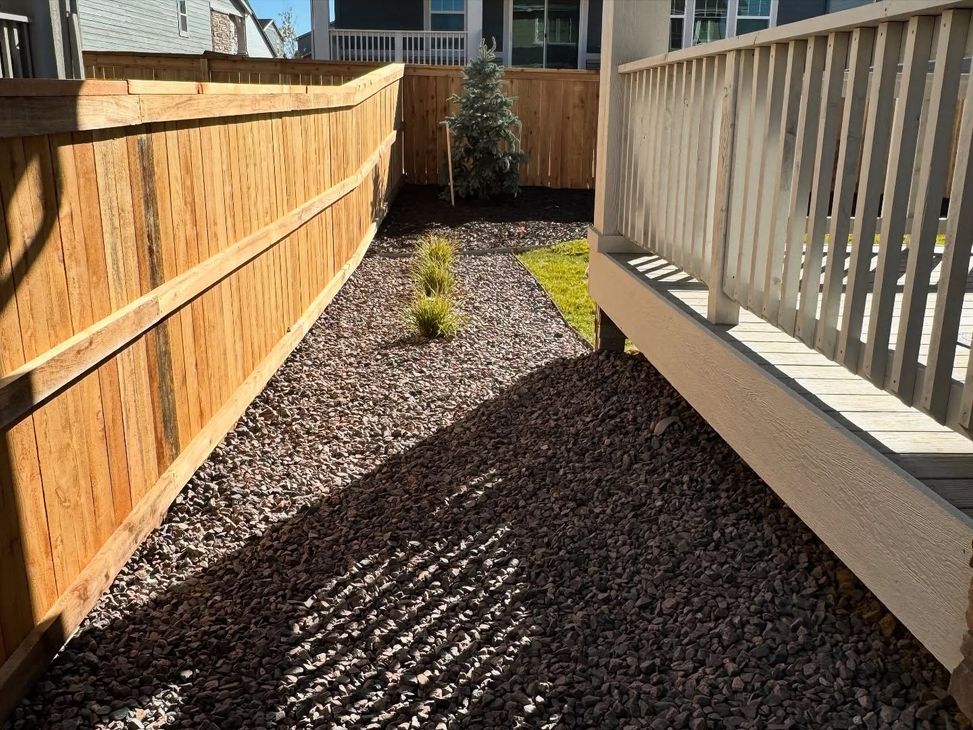 A gravel side yard featuring a wooden fence on the left and a porch railing on the right, with small shrubs in the center.