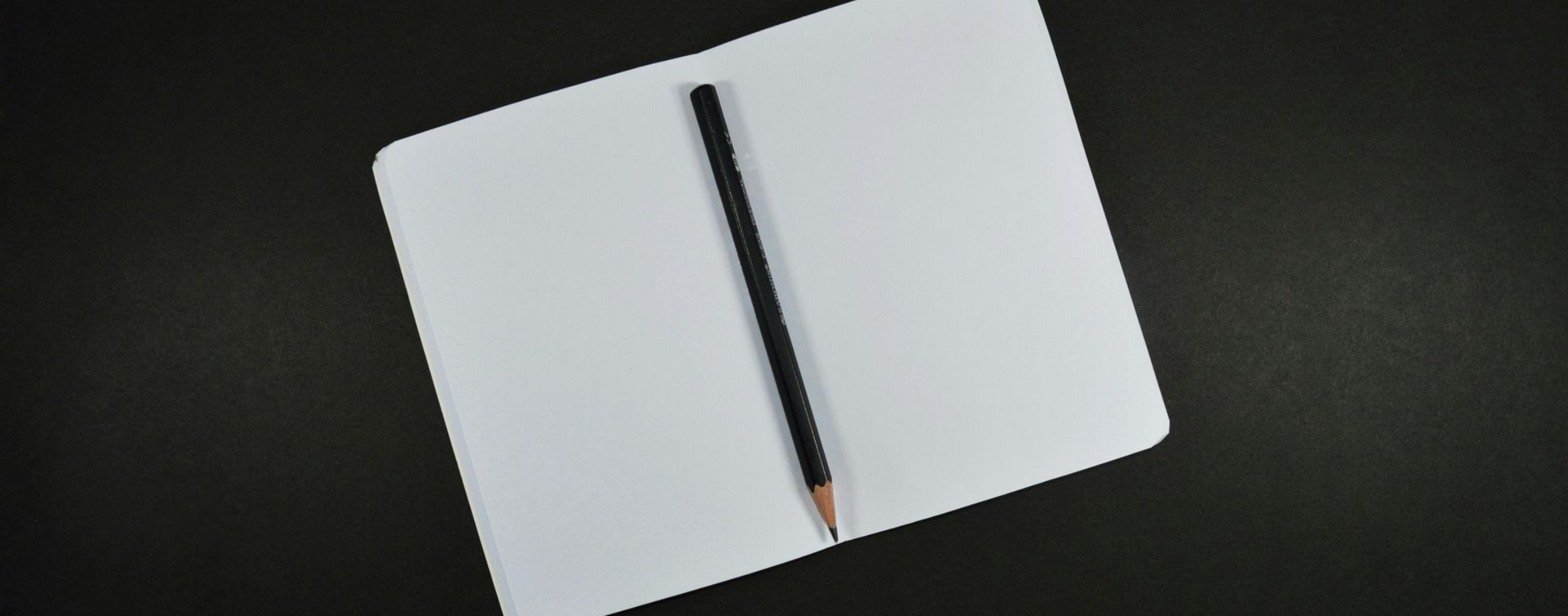 Open white notebook with a black pencil lying across the open pages on a dark surface.
