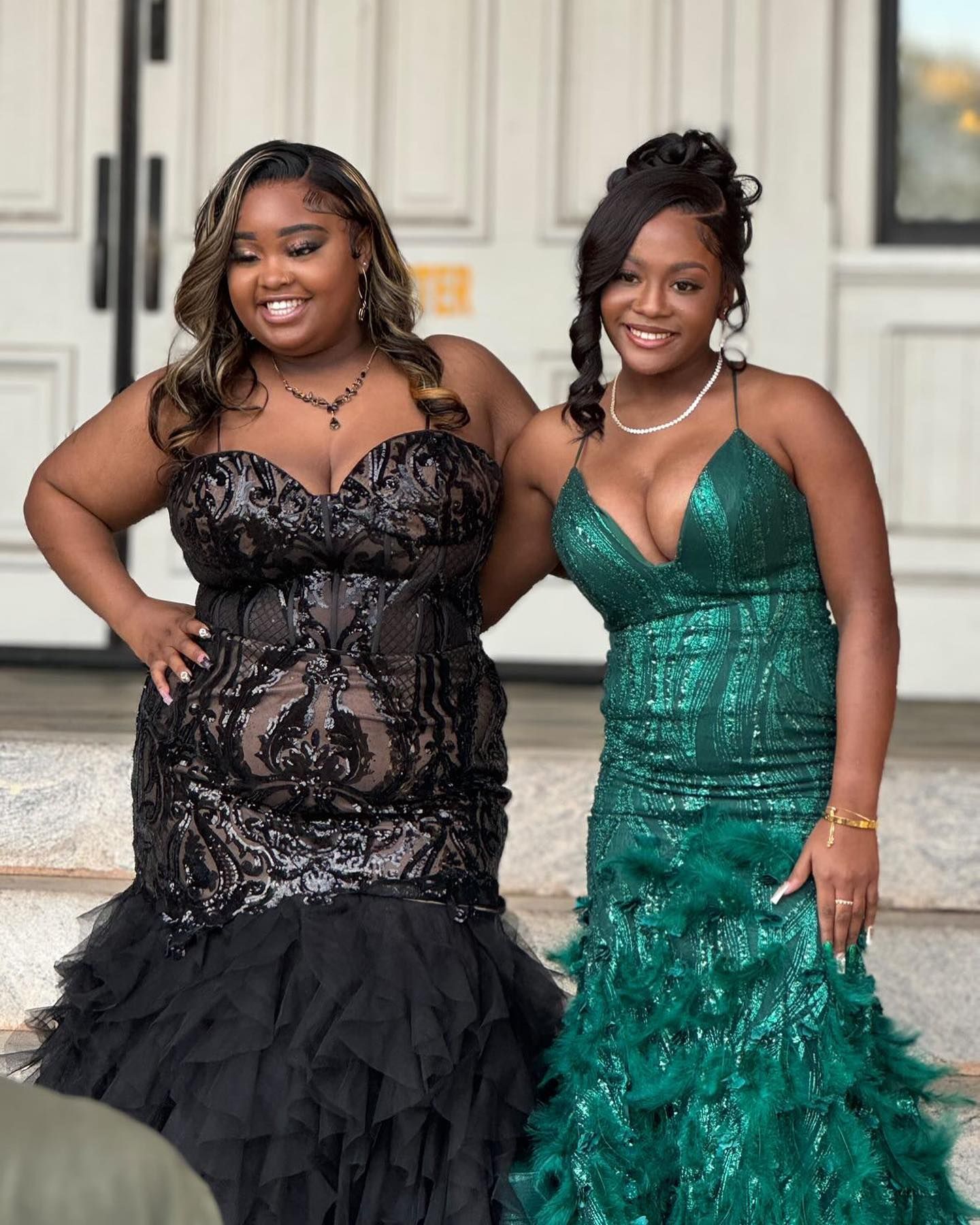 Two brides in white dresses smile at each other inside a greenhouse, holding bouquets.