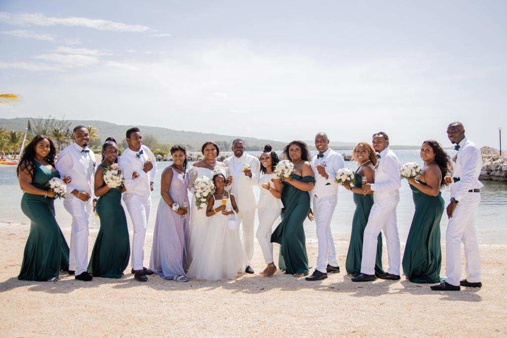 Wedding party on beach: bride, groom, bridesmaids in green and lavender, groomsmen in white suits.