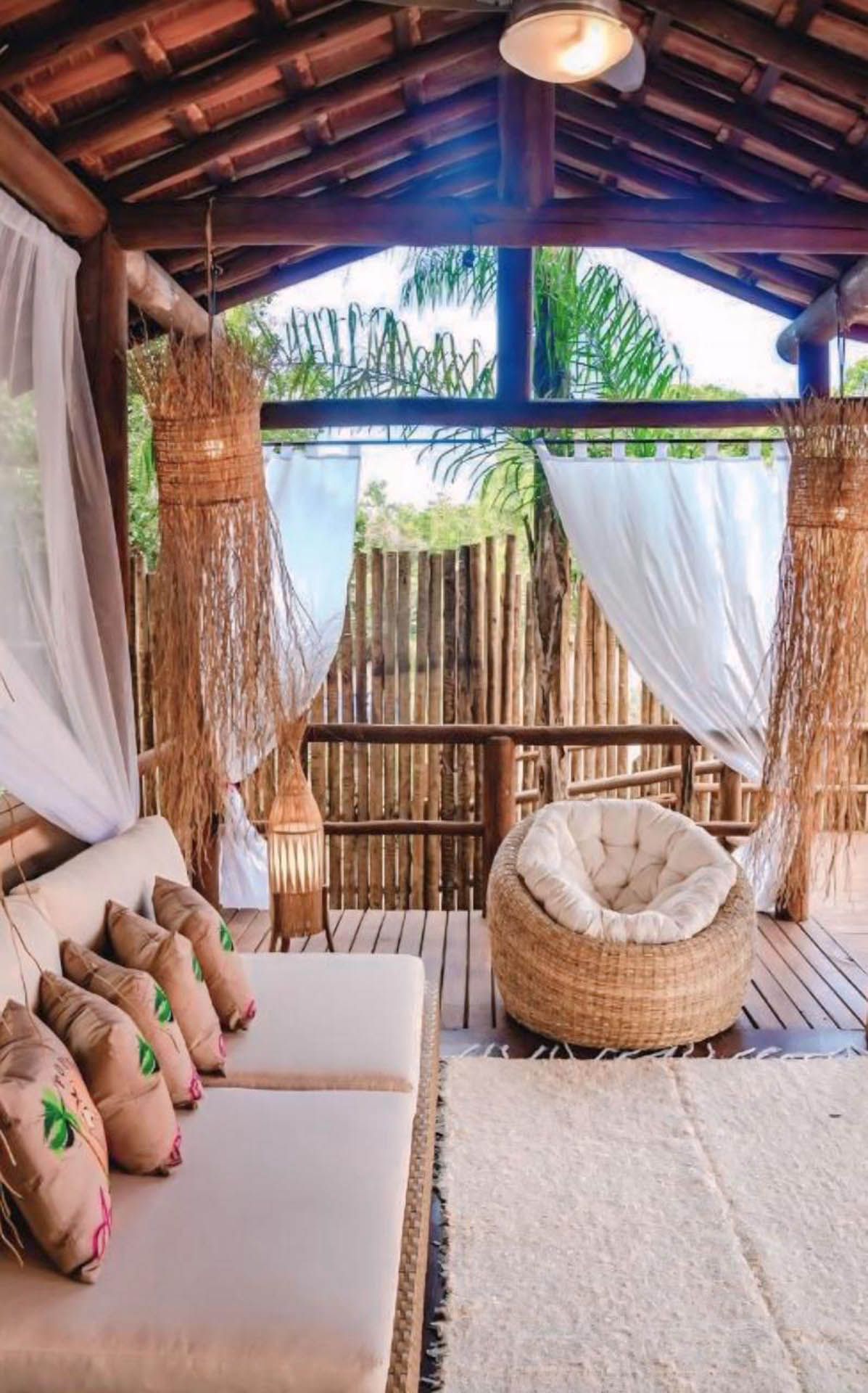 Aconchegante pátio boho com cadeira de vime, sofá bege, cortinas translúcidas e paredes de bambu sob um telhado de madeira.