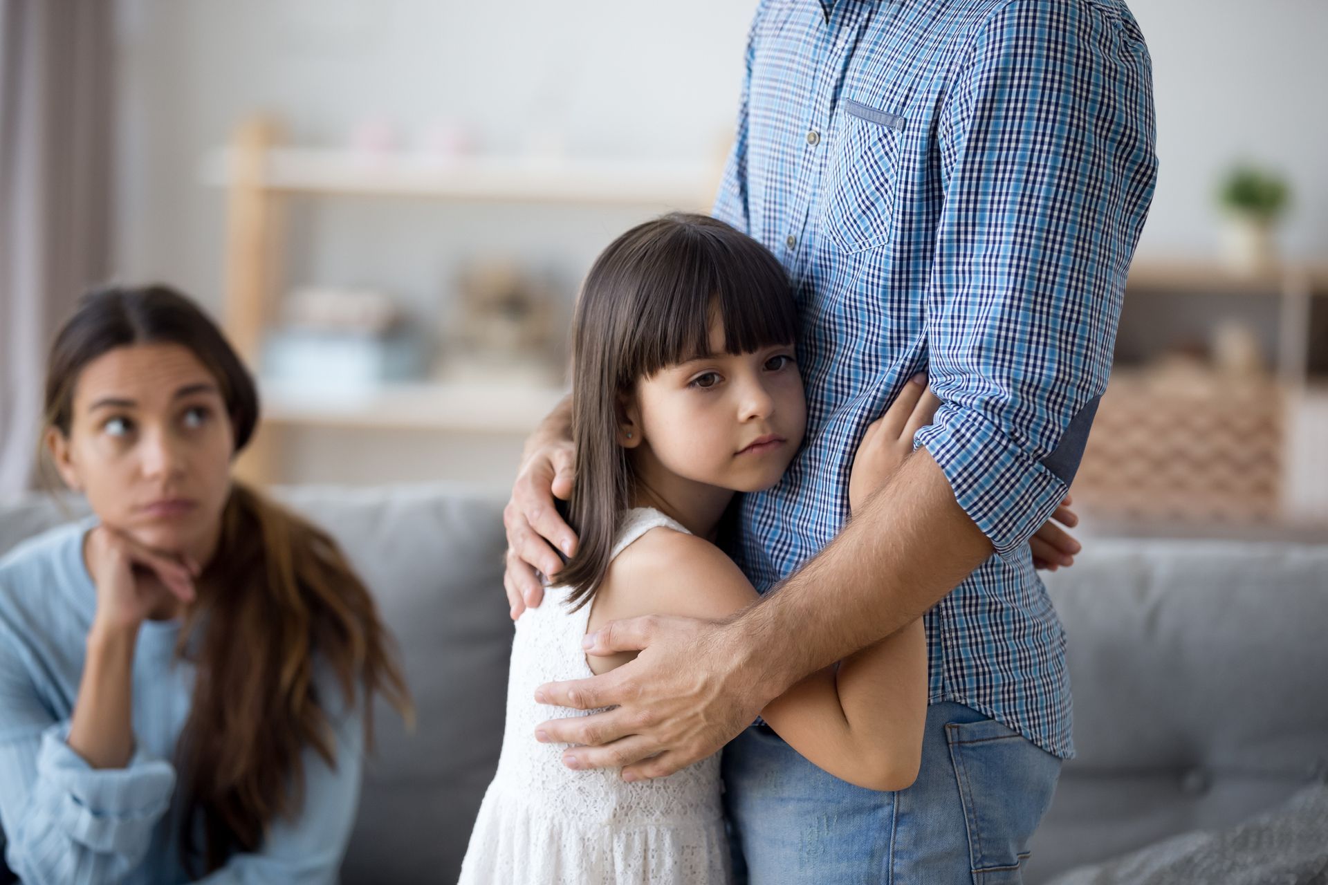 A child hugs a man while a woman looks on with a concerned expression. Indoors, living room.