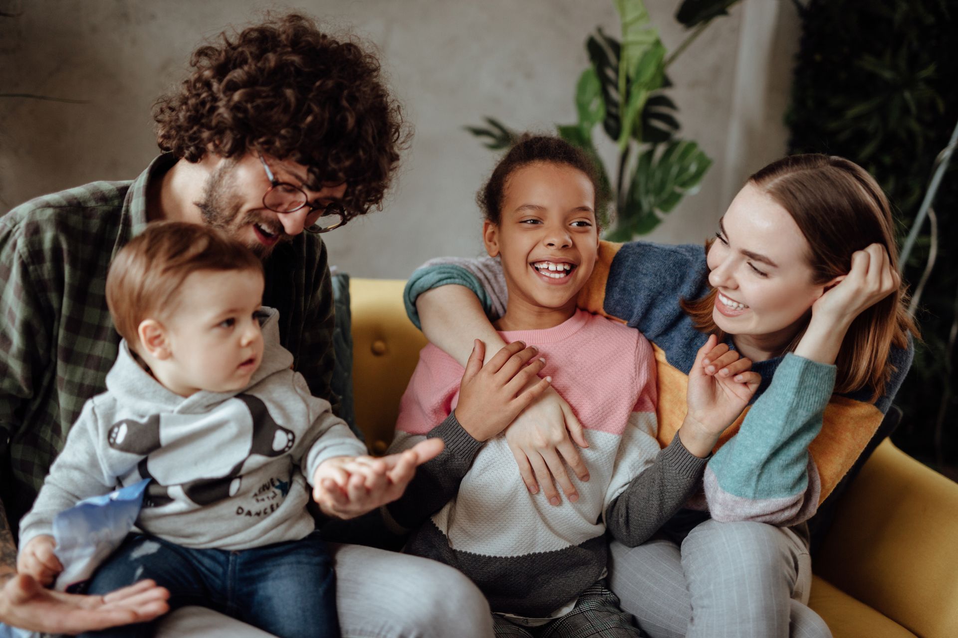 Family of four on a couch, laughing. A woman embraces a girl, while a man holds a baby.
