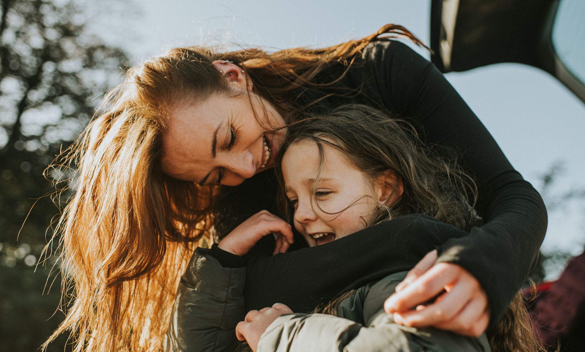 Woman with long hair hugging a laughing child outdoors.