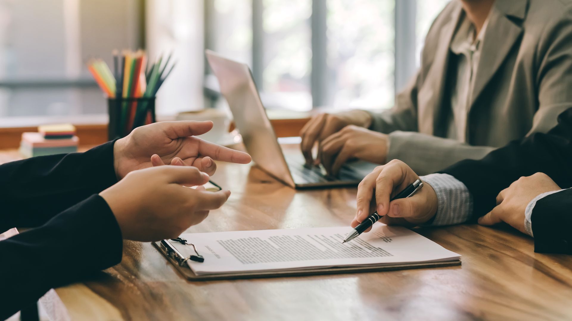 People in business attire review a document at a wooden table, one using a laptop.