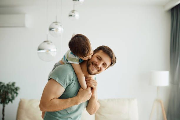 Father smiling with child on his shoulders, kissing his cheek; home interior.