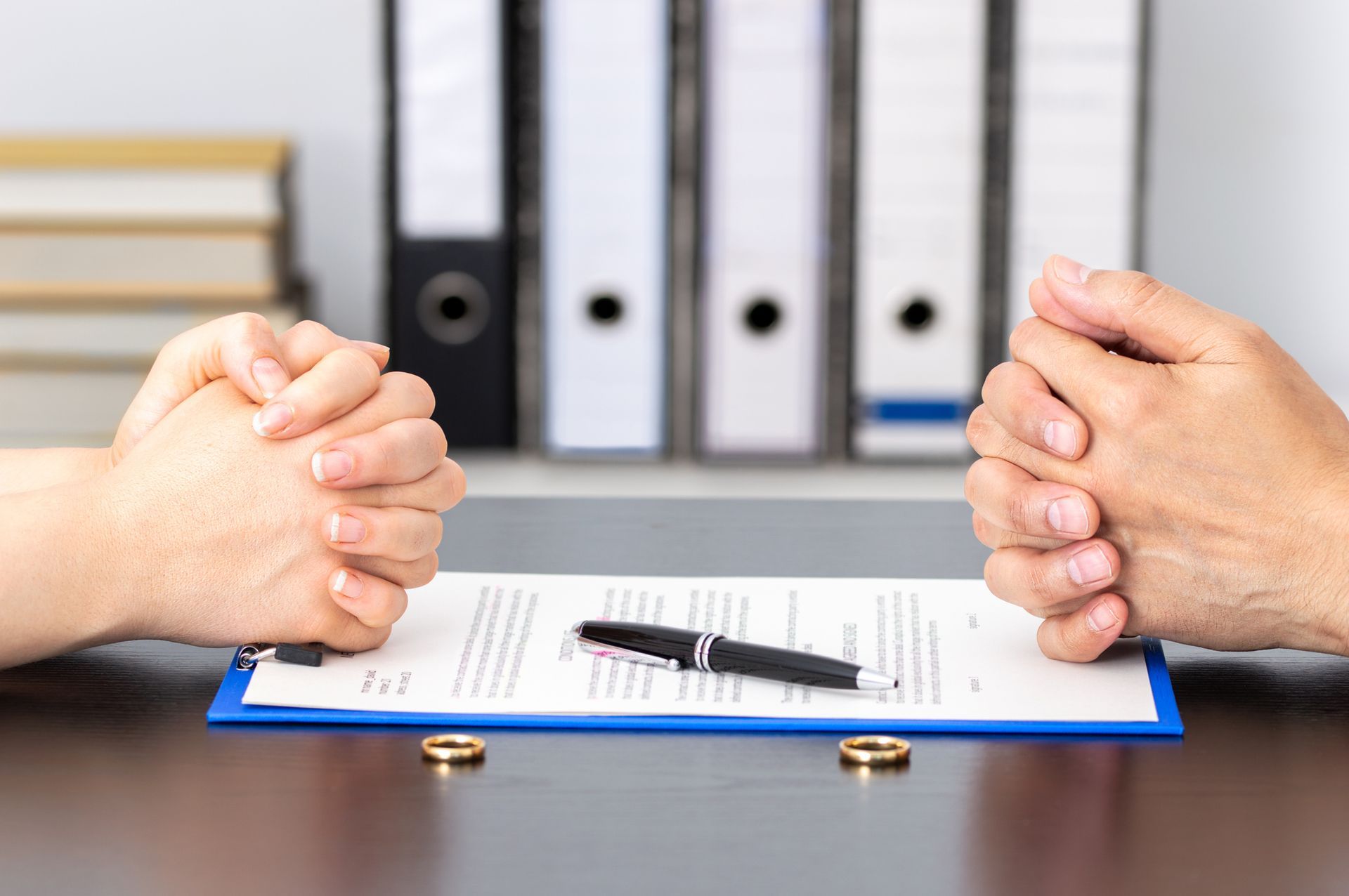Hands clasped over divorce papers and rings on a desk, symbolizing separation.