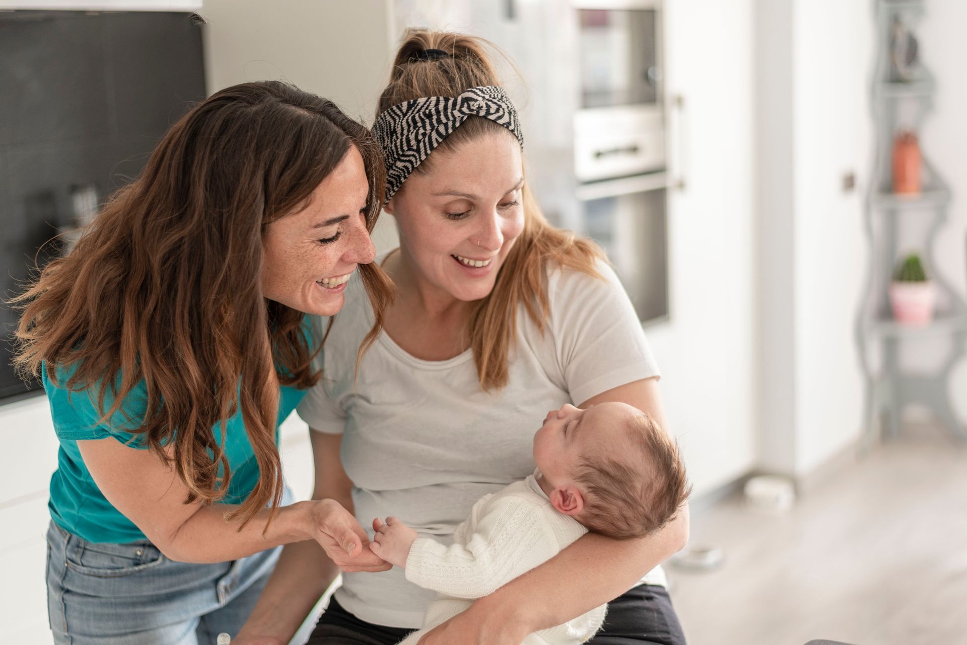 Two women, smiling, one holding a baby. Kitchen setting, bright, white shirt, teal top, baby in white.