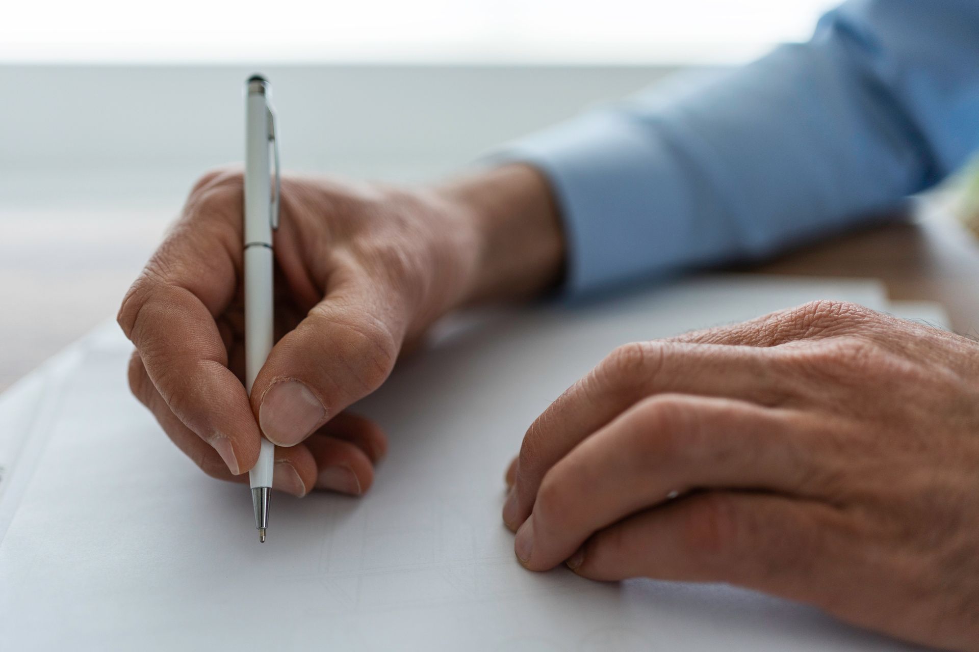 Person's hands writing on a white paper with a silver pen, wearing a blue shirt.