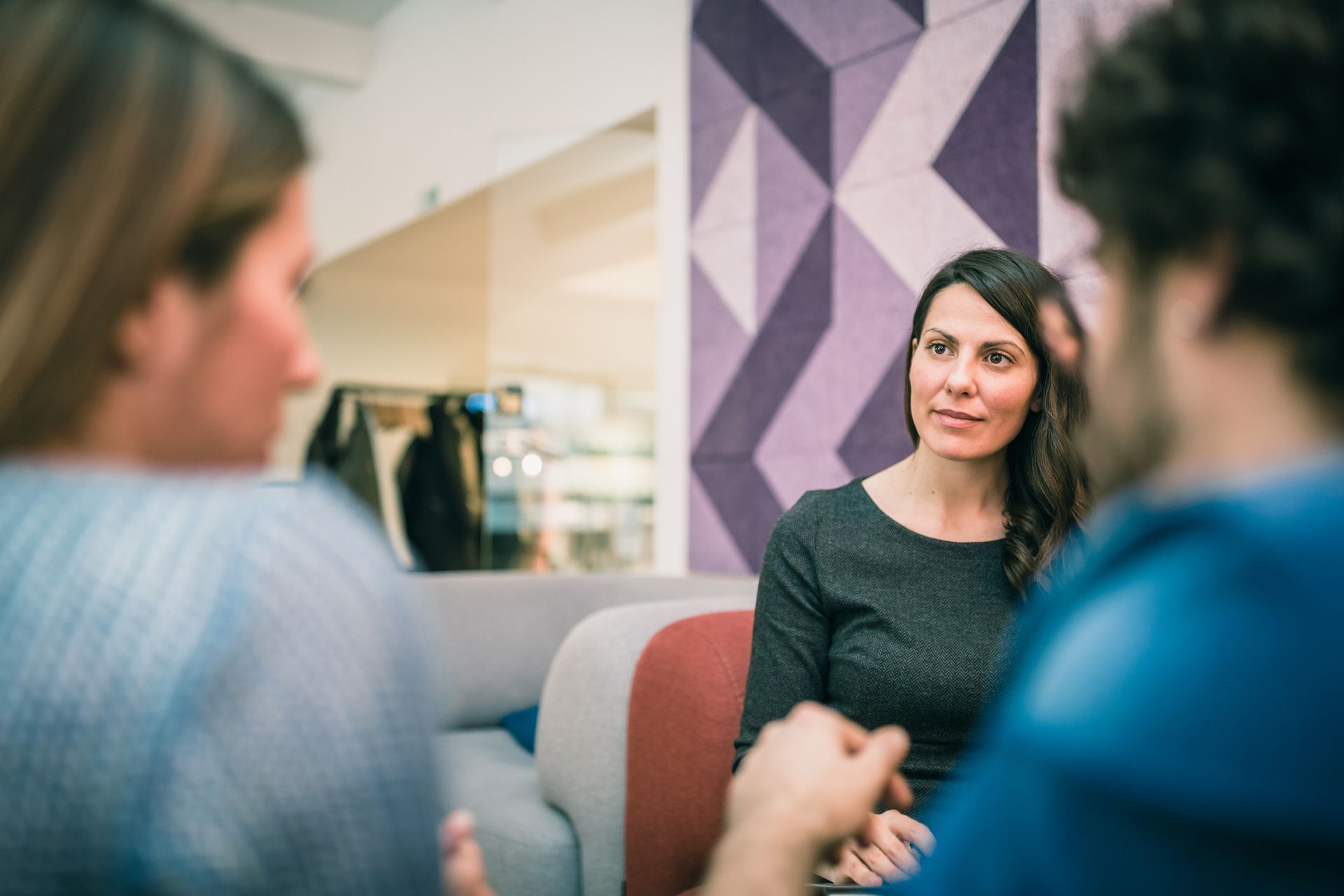 Woman with long hair listens attentively to two people in a modern office setting; geometric wall in background.