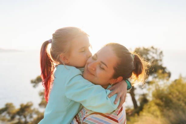 A young girl hugs and kisses her mother outdoors, both smiling in the sunlight.
