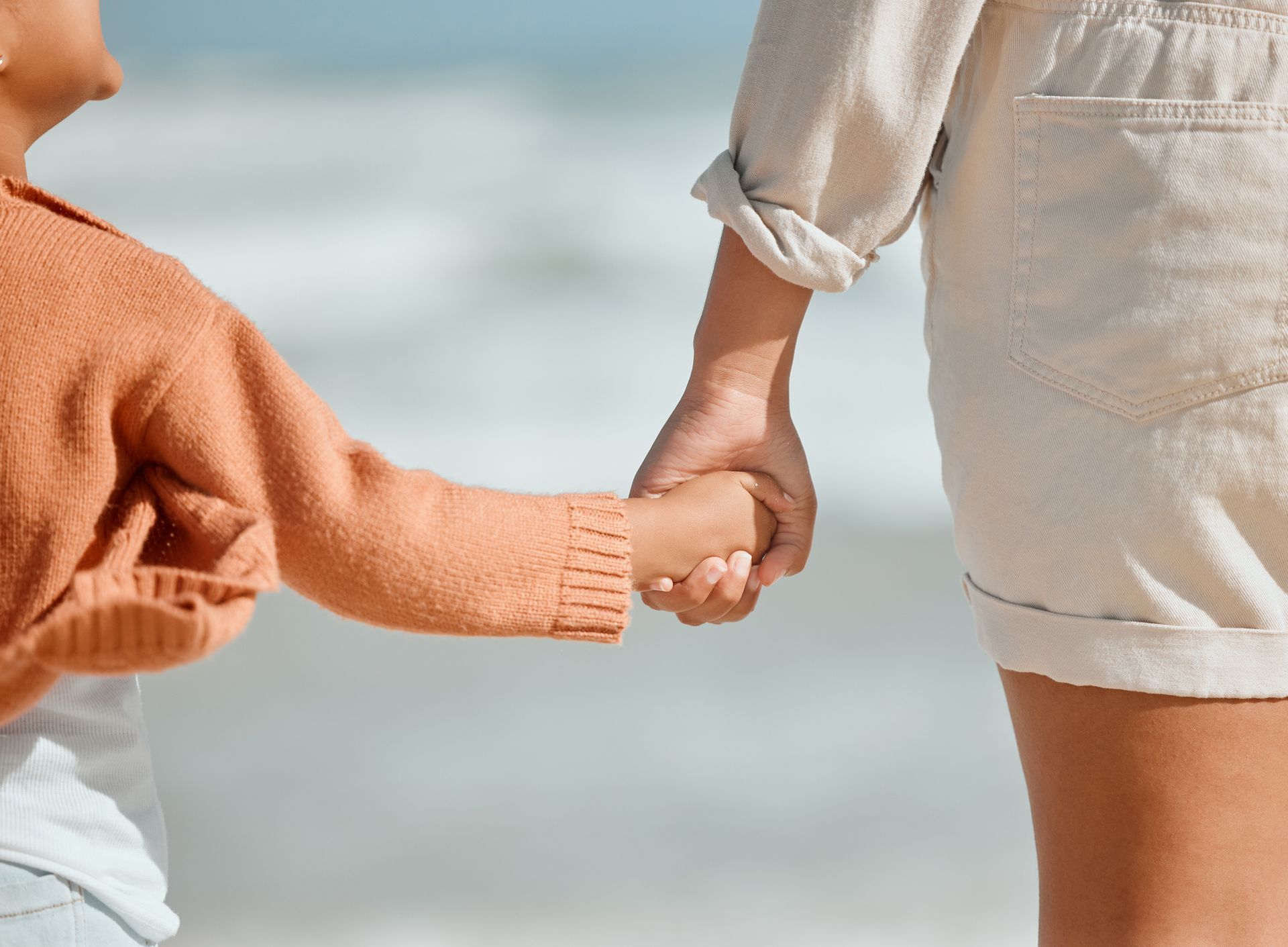 Child holding an adult's hand on a beach. The child wears an orange sweater.