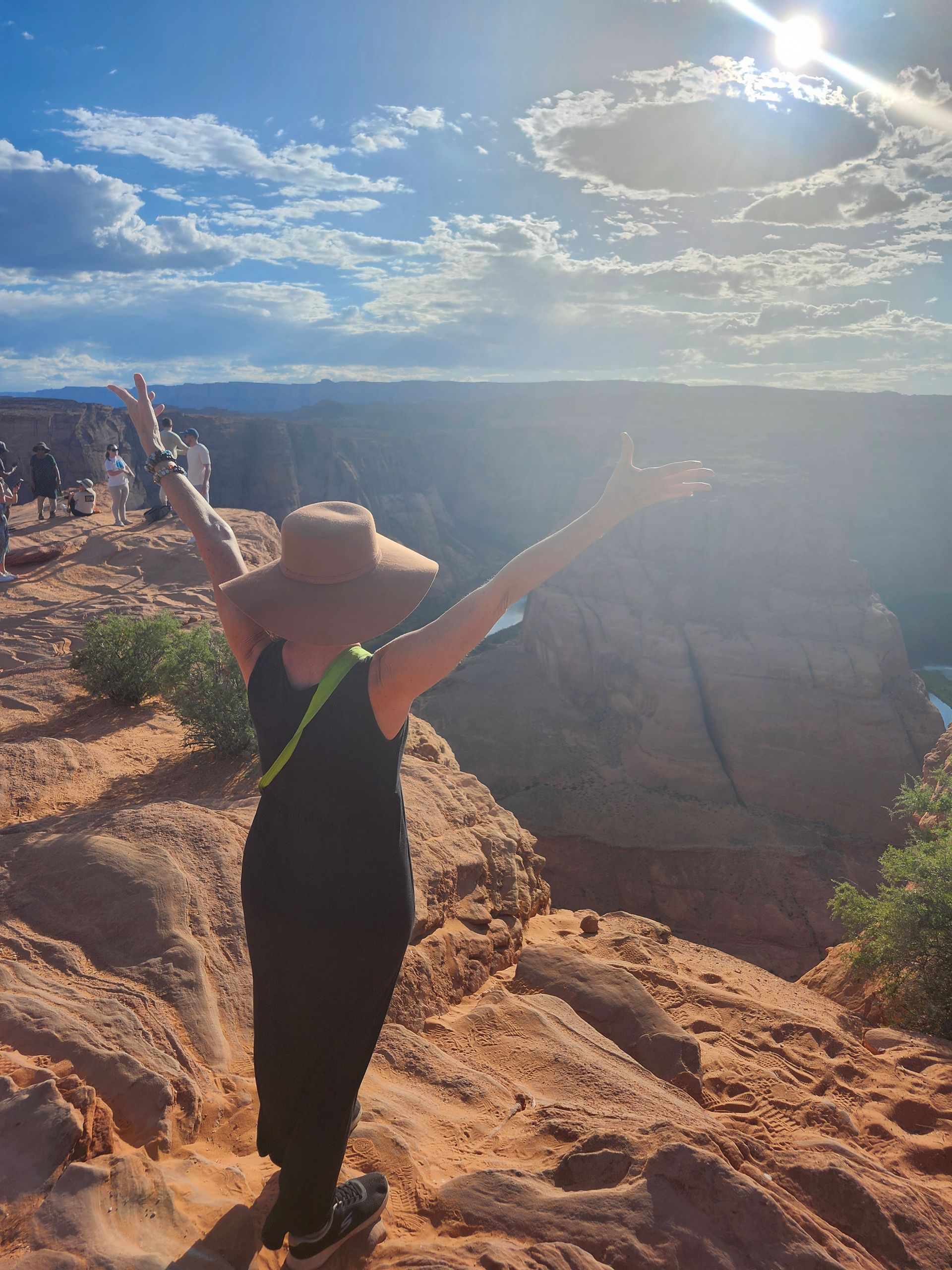 A woman in a hat is standing on top of a rocky cliff with her arms outstretched.