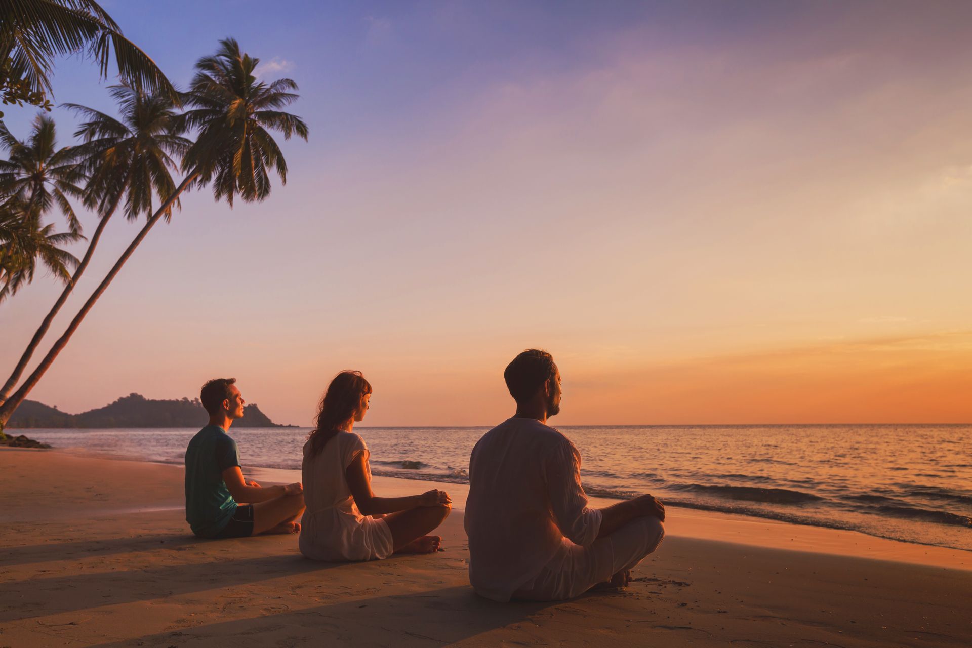Three people are sitting in a lotus position on the beach at sunset.