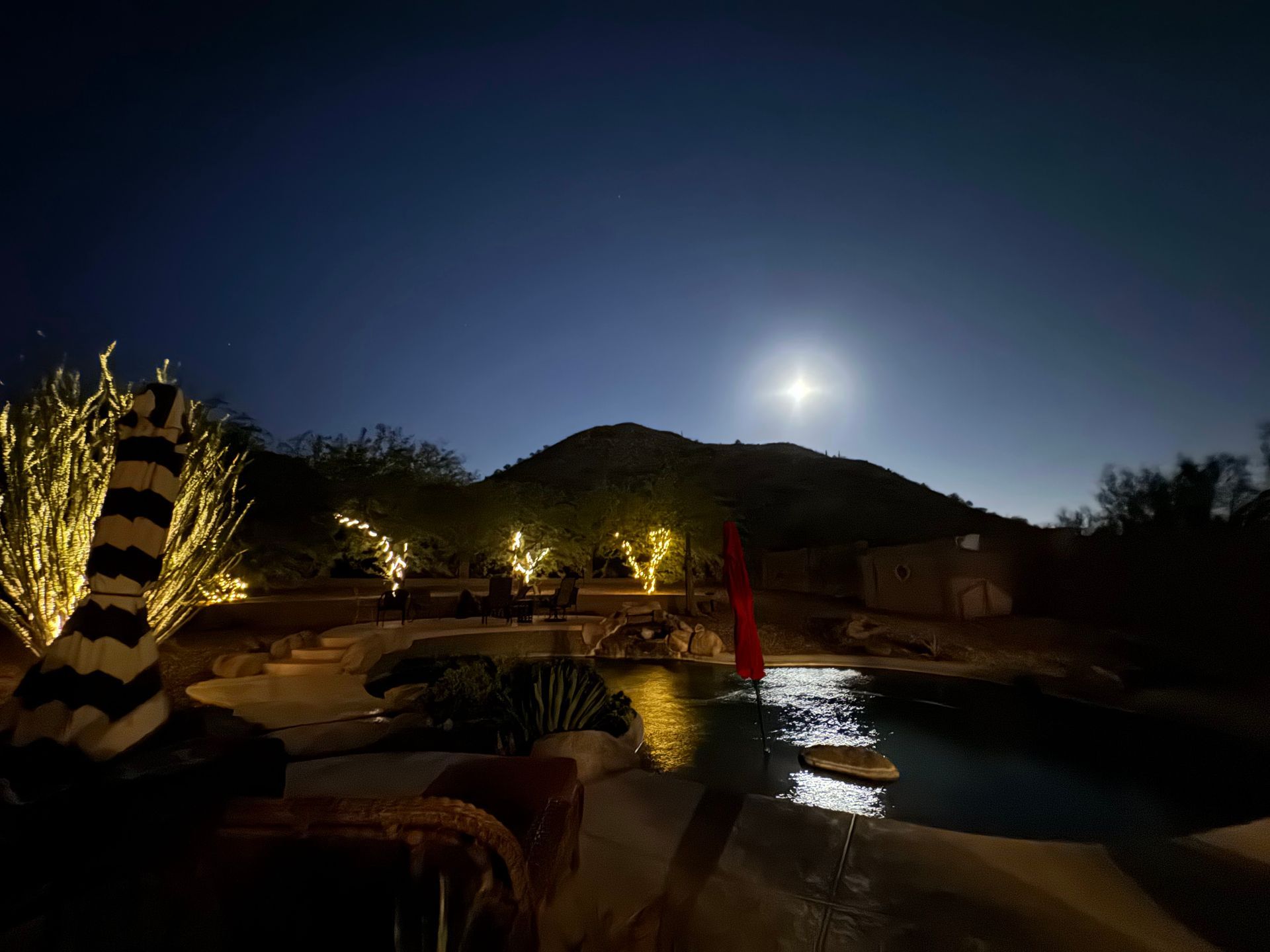 A swimming pool is lit up at night with a full moon in the background.