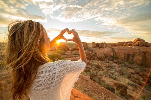 A woman is making a heart shape with her hands in front of the sun.