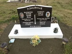 Black granite headstone with photos, names, and dates; set on a white platform with small floral arrangements — Mackay Monumental Masons In Glenella, QLD