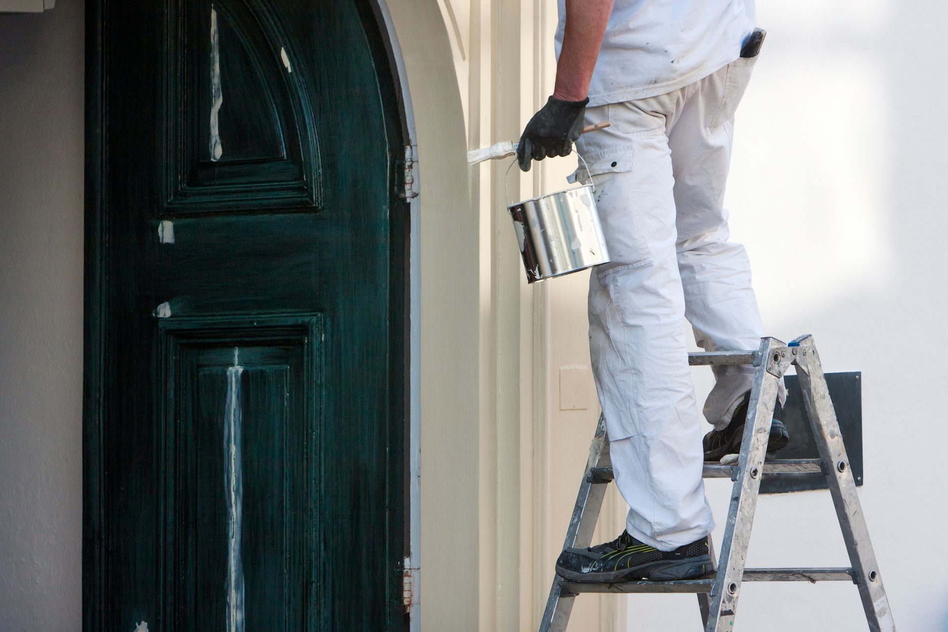 Person painting green door trim while standing on a ladder. White pants, black gloves, paint can.
