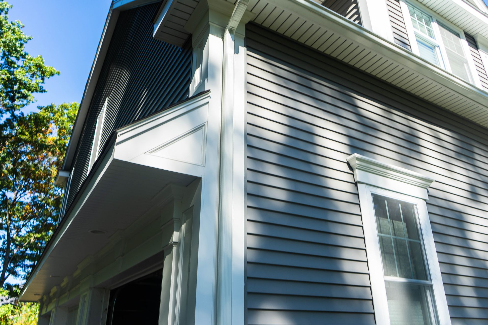Corner of a two-story gray house with white trim, garage, and a window against a blue sky.