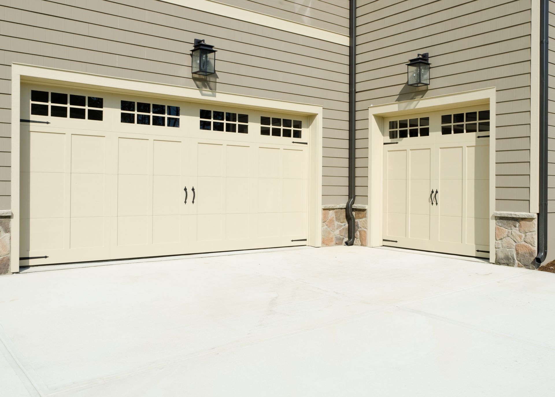 Two cream-colored garage doors with windows above, on a concrete driveway, against a beige siding.