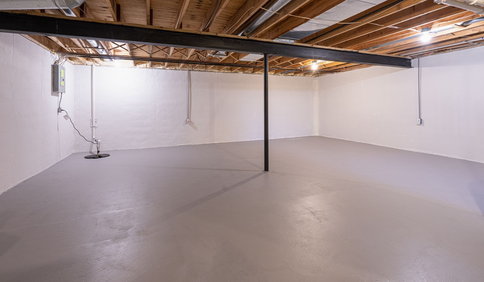 Empty basement with gray painted floor and white walls. Exposed ceiling beams and black support beam.