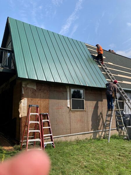 Construction workers installing green metal roofing on a building, using ladders. Sunny day.