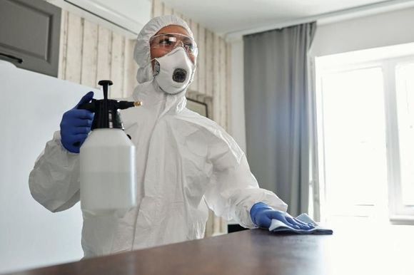 Una persona con indosso una tuta protettiva bianca, una maschera e dei guanti utilizza un flacone spray per disinfettare una superficie in una stanza.