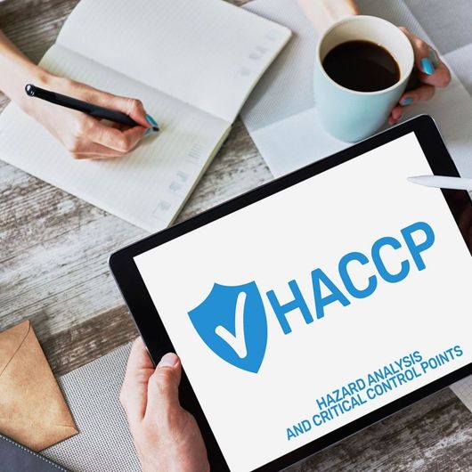 Su una scrivania, accanto a un quaderno, una penna e una tazza di caffè, lo schermo di un tablet mostra il logo HACCP blu e del testo.