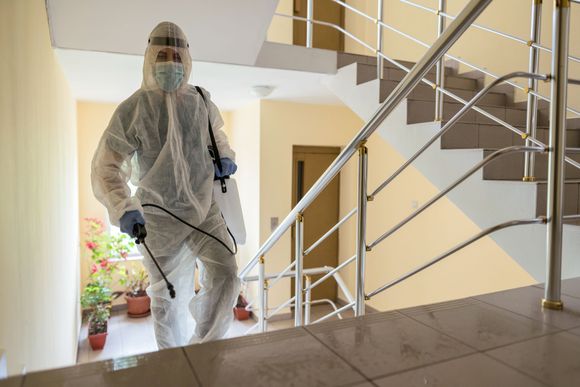 Una persona con indumenti protettivi e mascherina utilizza uno spruzzatore per igienizzare una scala interna e il pianerottolo.