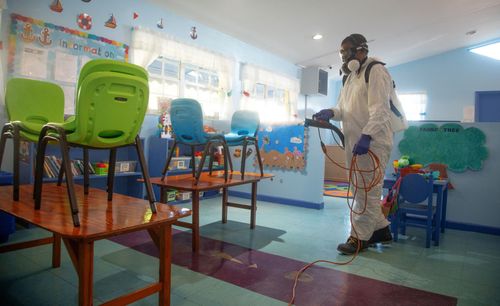 Una persona con tuta protettiva e mascherina spruzza disinfettante in un'aula con sedie impilate sui tavoli.