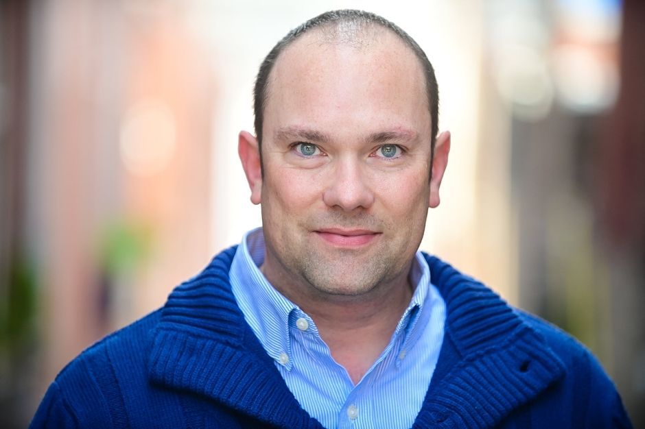 Portrait of a smiling person in a blue sweater and light blue shirt outdoors with a blurred background