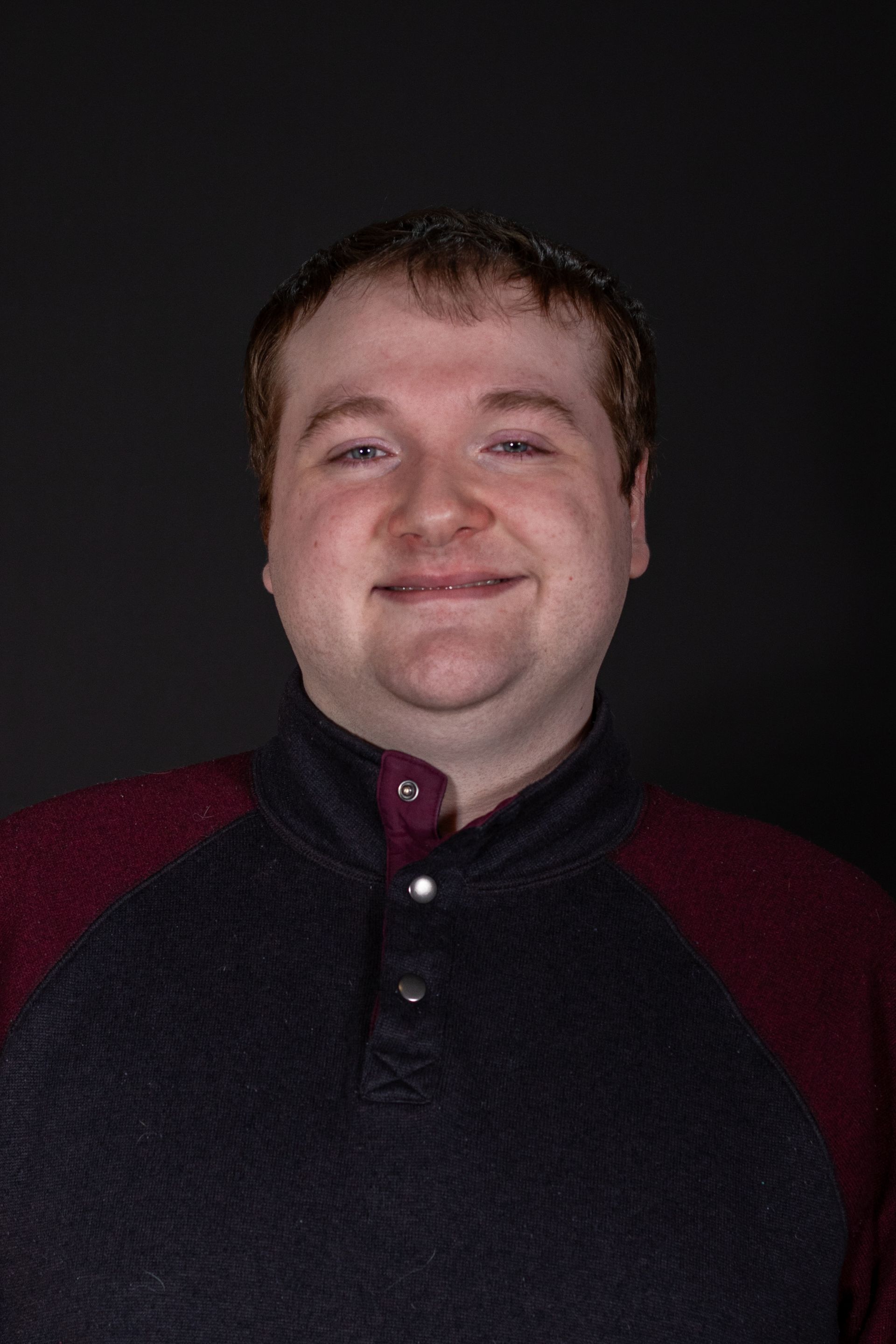 Man in a burgundy and navy shirt smiles against a dark background.