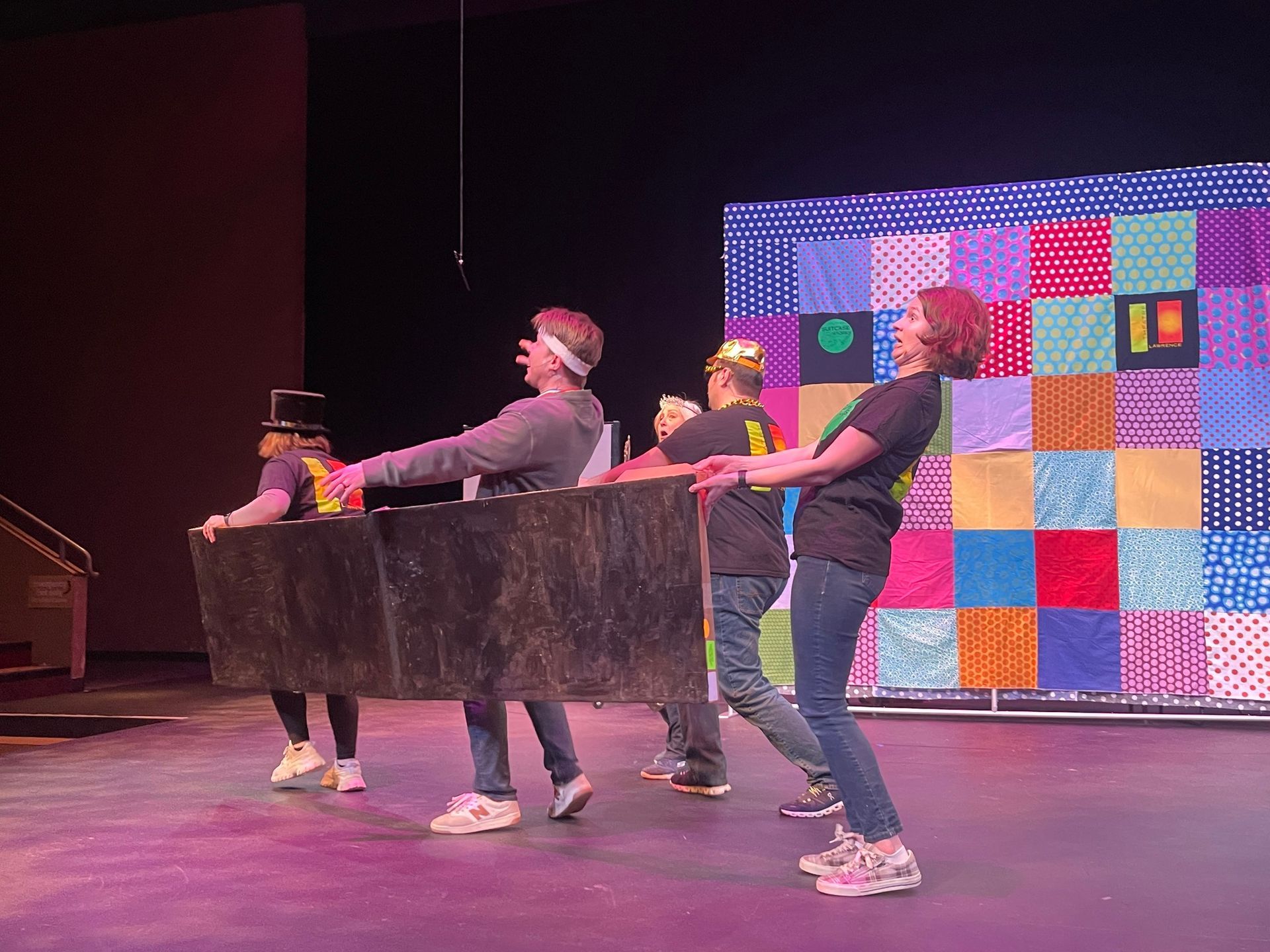 People on stage, pulling a prop. The prop appears to be a long, black, rectangular shape. Colorful backdrop.