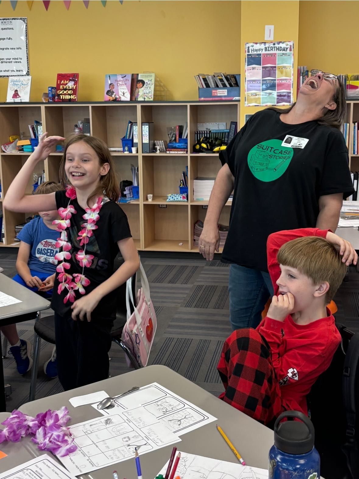 Girl in lei laughs with teacher and boy at desk in classroom.