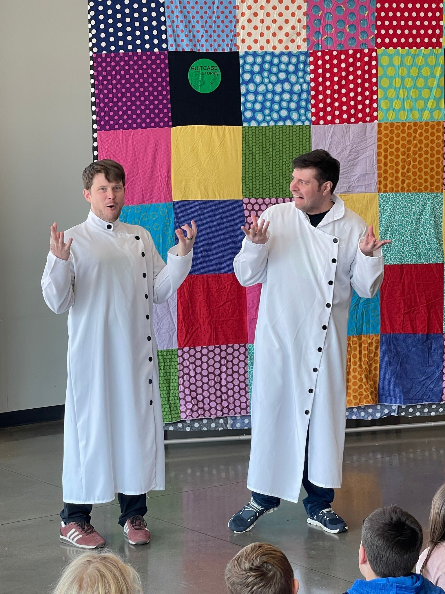 Two people in white coats gesturing in front of a colorful quilt.