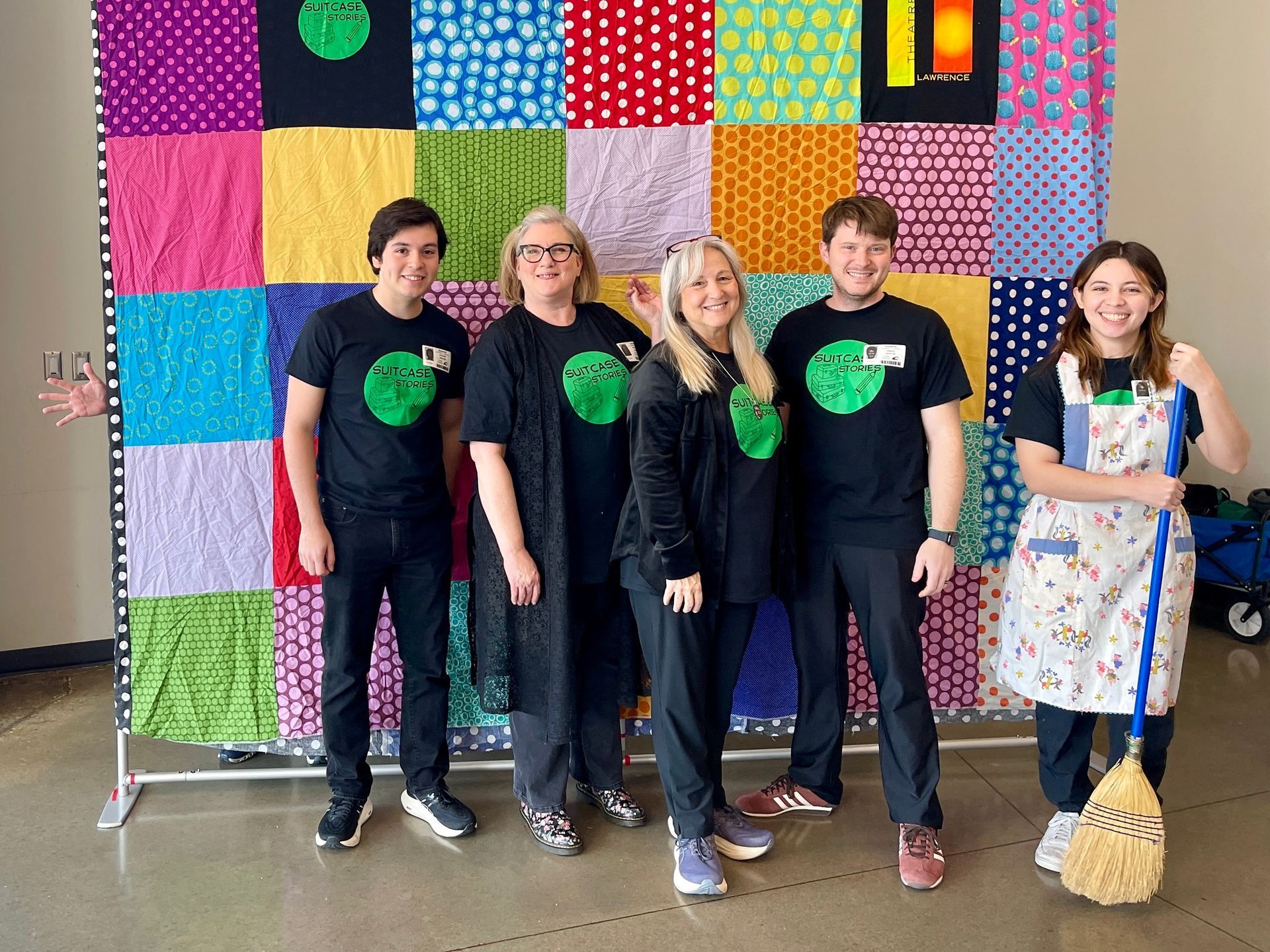Five people in black shirts pose in front of a colorful quilt. One holds a broom.