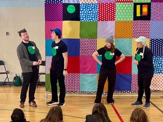 Five people in black shirts pose in front of a colorful patchwork quilt; one holds a broom.