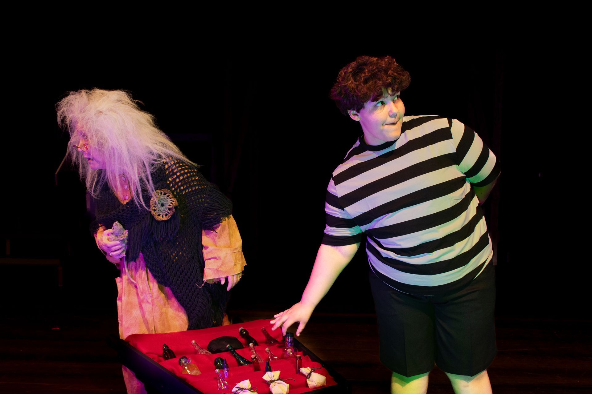 Two people at a red-lit table, one reaching toward objects while the other looks down with a white wig.