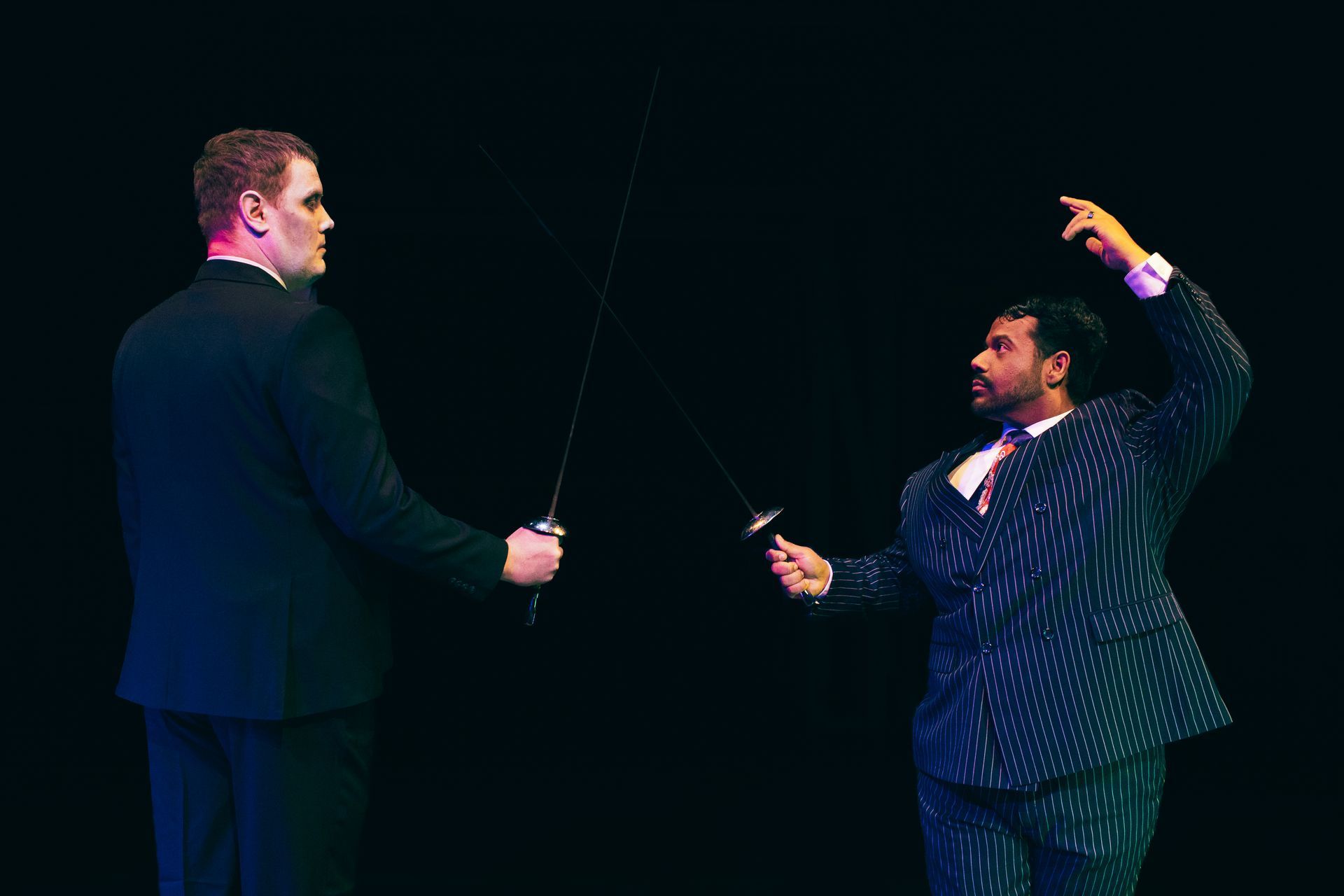 Two men in suits facing each other on a dark stage, each holding a sword, one with an arm raised