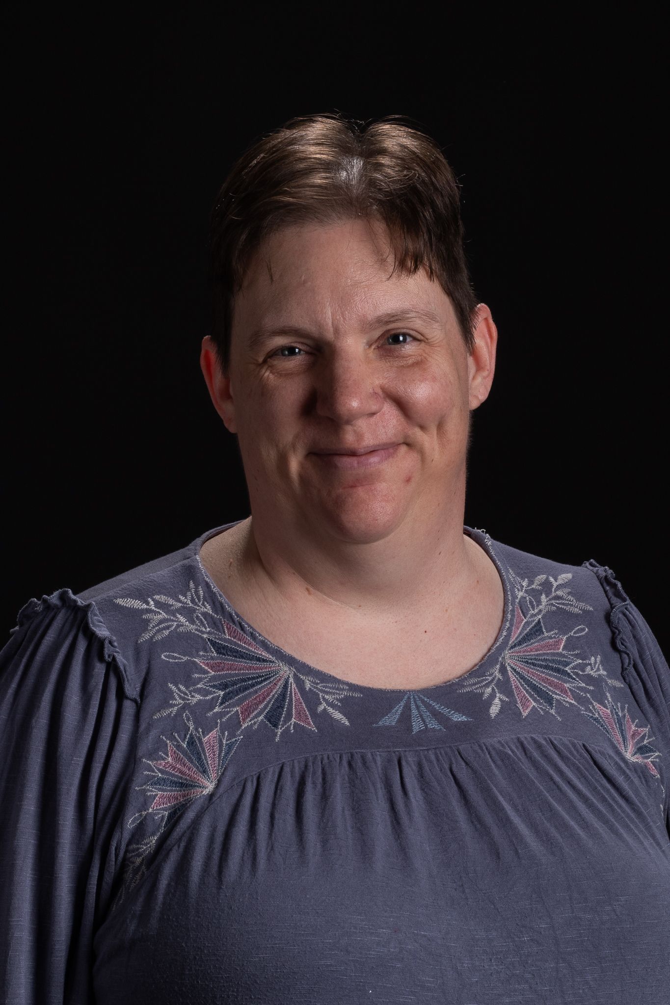 Woman with short hair smiles, wearing a purple shirt, in front of a dark background.