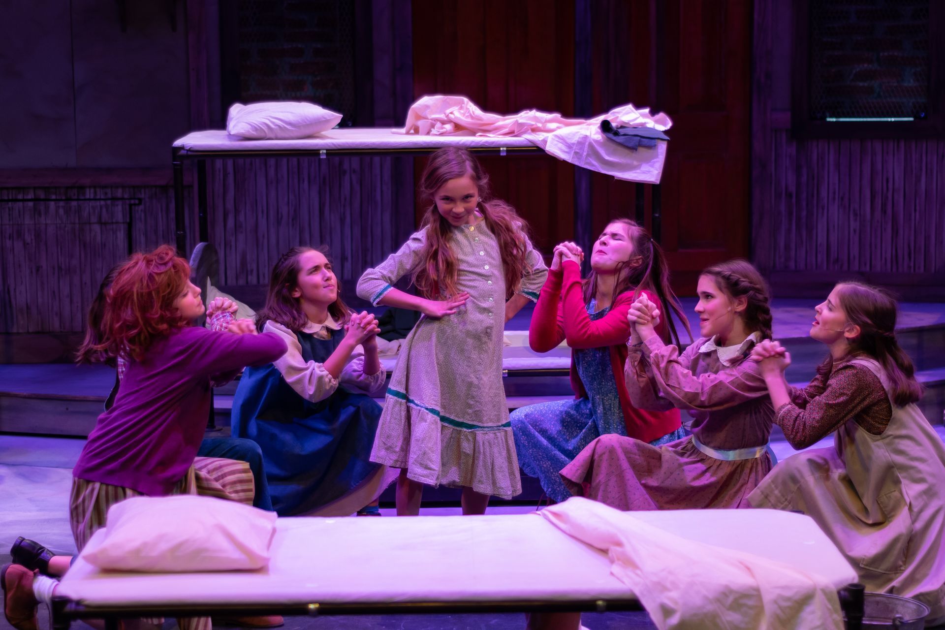A stage production scene: seven young women in period clothing praying around a bed.