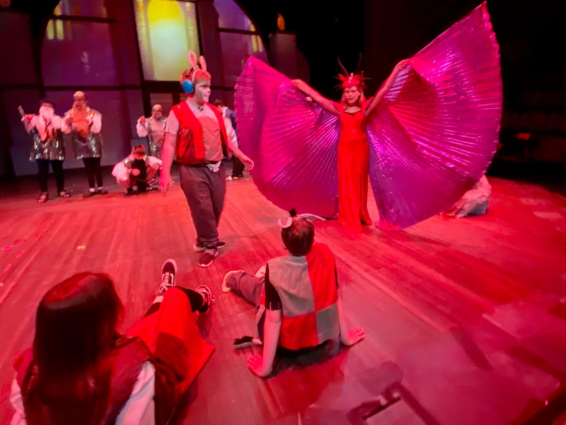 A group of people are performing on a stage with pink wings.