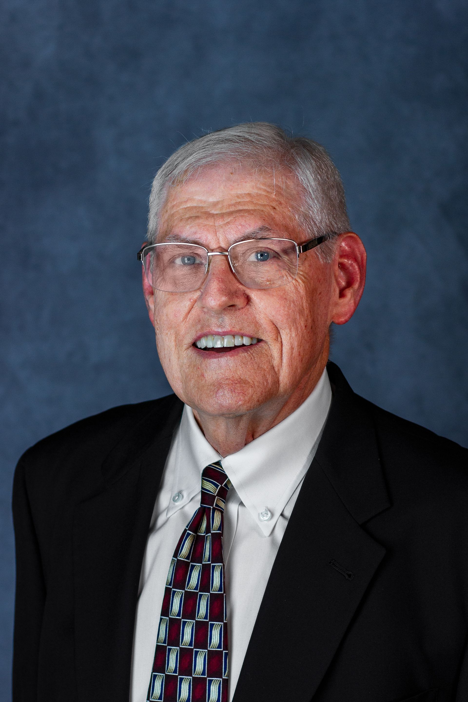 An elderly man in a black suit and tie smiles, wearing glasses against a dark blue background.