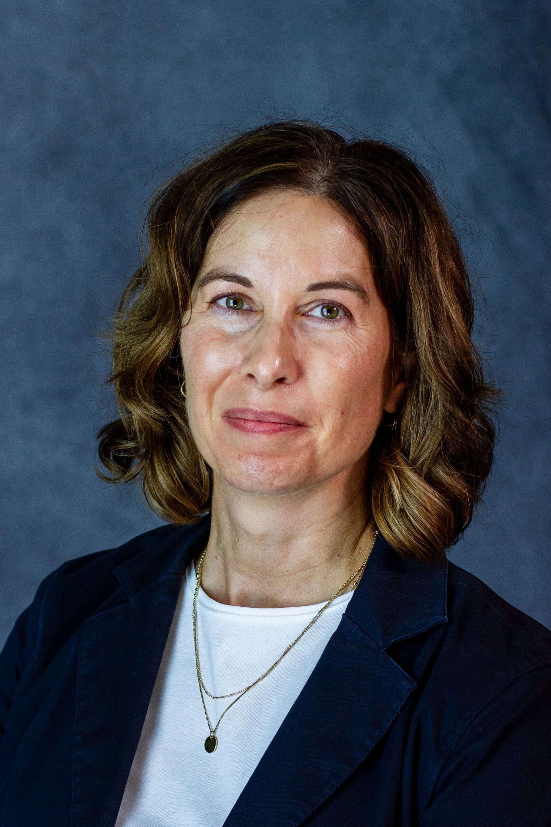 Woman with brown hair in a navy blazer smiles at the camera. She wears a white shirt and necklace.