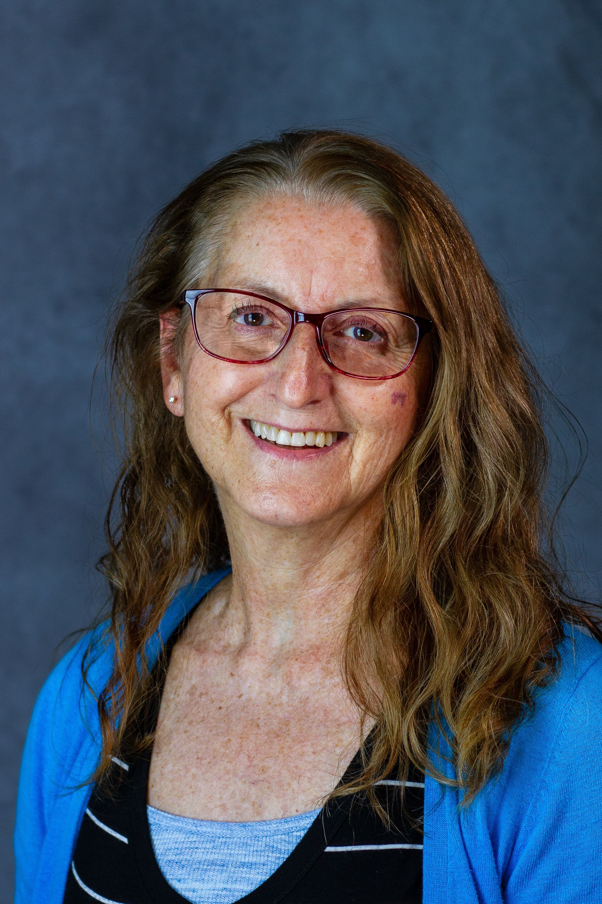 Woman with glasses smiling, wearing a blue cardigan over a black and white striped top, against a dark gray backdrop.