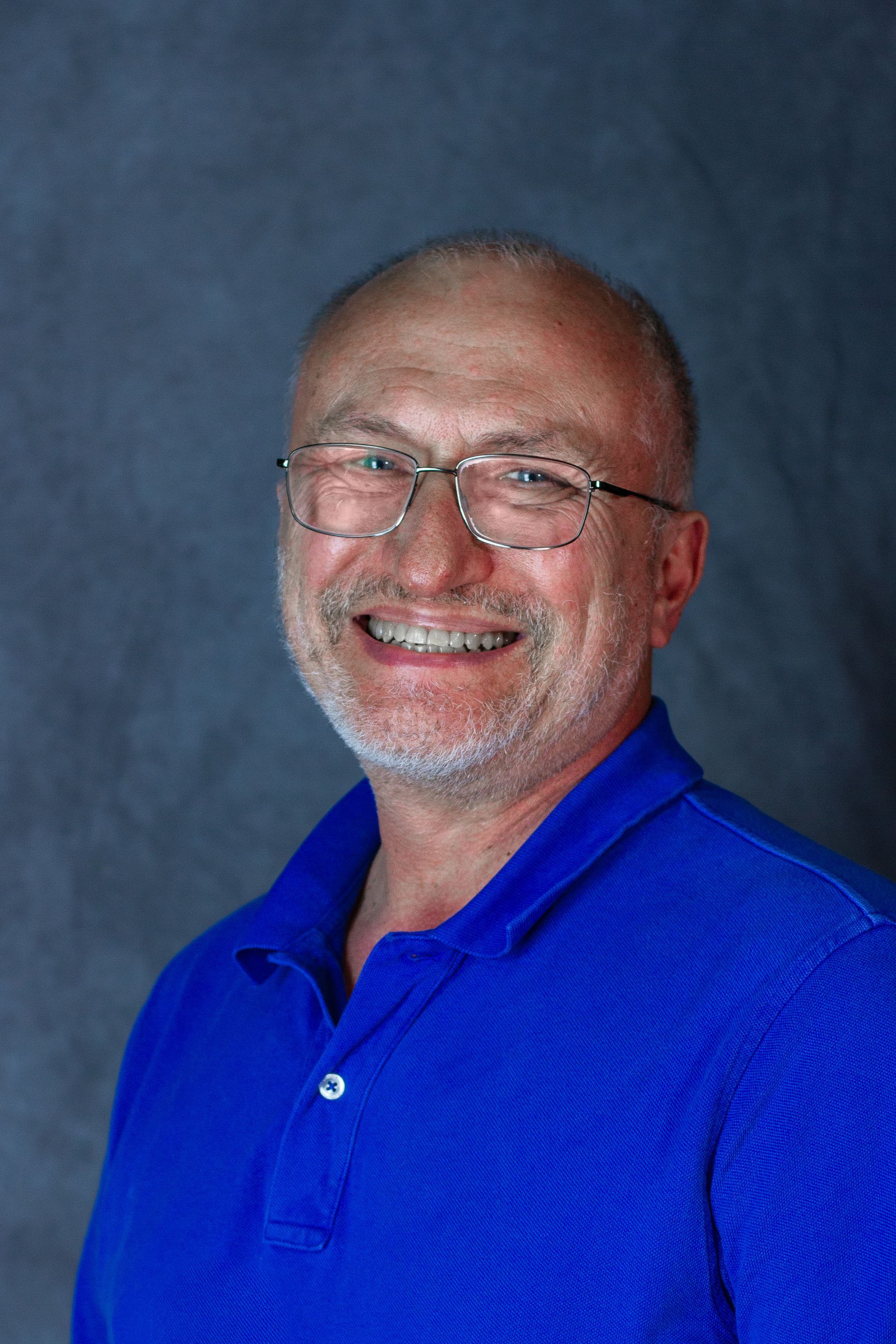 Smiling man with glasses, blue shirt, against a blue-gray backdrop.
