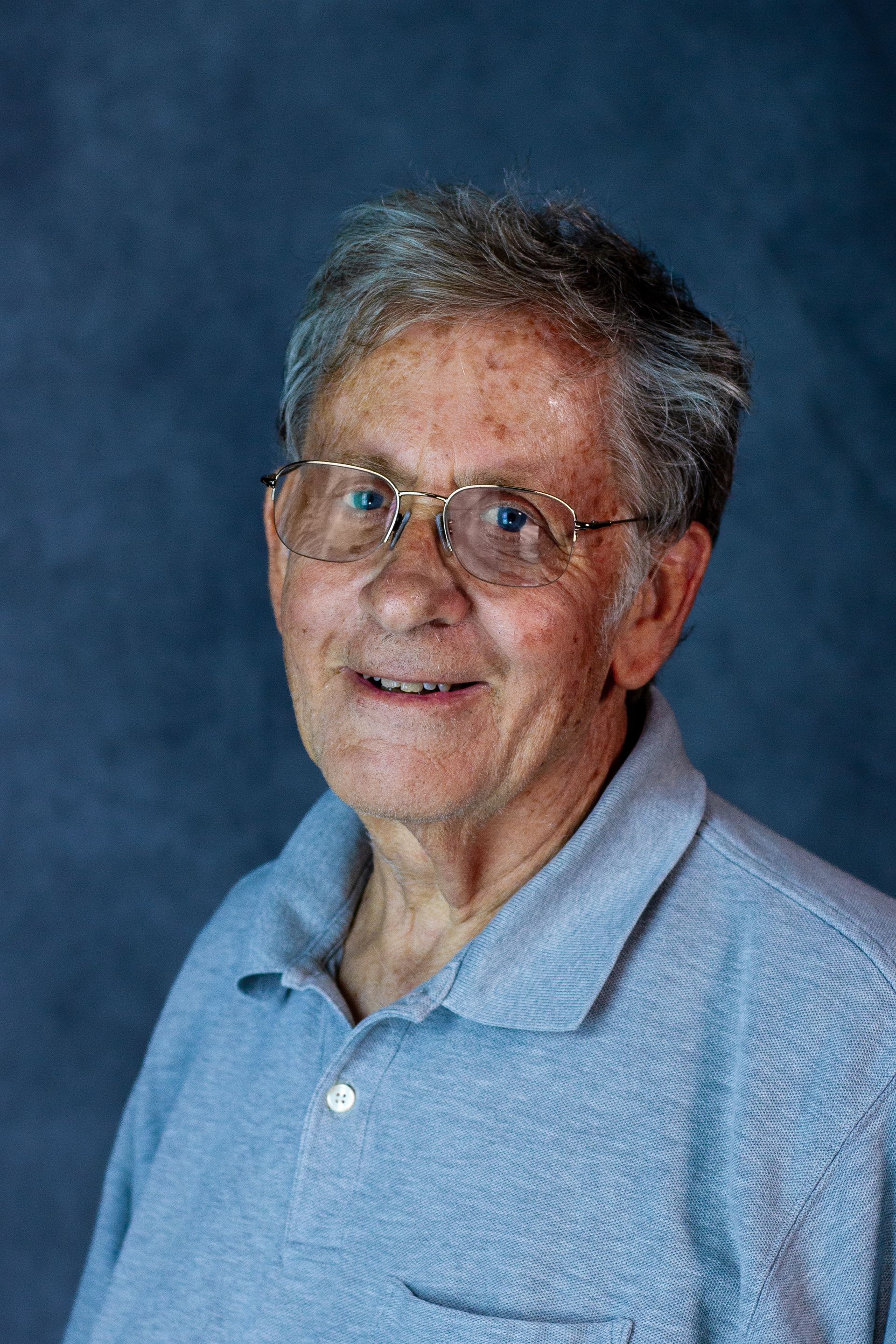 Elderly man wearing glasses and a blue polo shirt smiling.