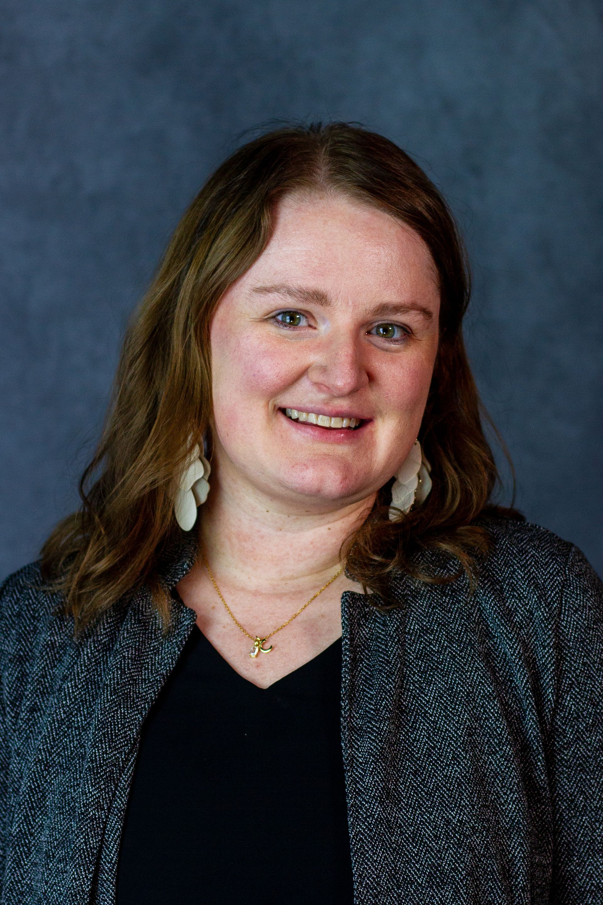 Woman with light skin smiles, wearing a black top, patterned jacket, and white earrings, posing in front of a blue background
